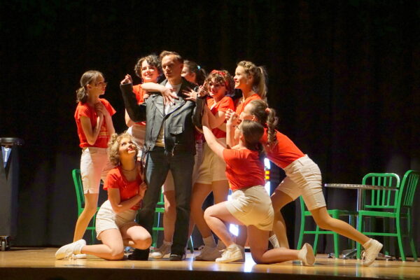 A group of young ladies dressed as carhops fawn on "The Fonz," who is in the middle in a leather jacket.