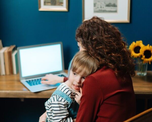 A mother works at the computer while her daughter rests on her shoulder.