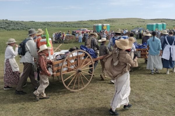 A group of young people dressed as pioneers pushes a handcart along a trail.