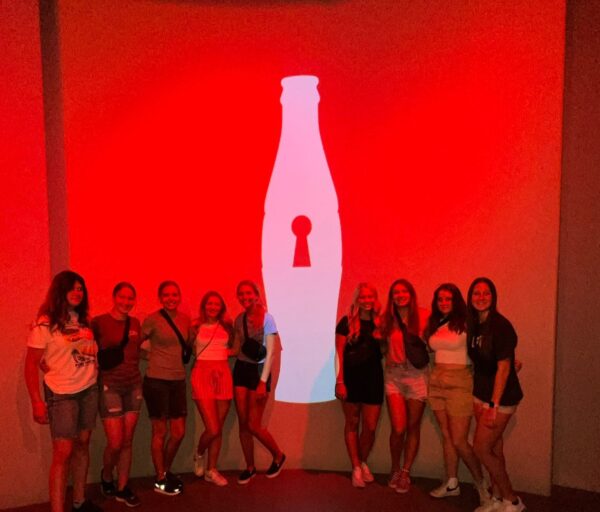 A group of girls next to a Coca-Cola painted on a red wall.