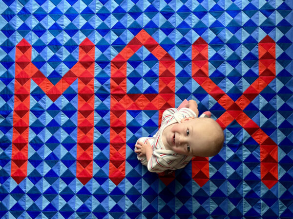 A baby sits on a quilt with the word "Max" in quilted squares.