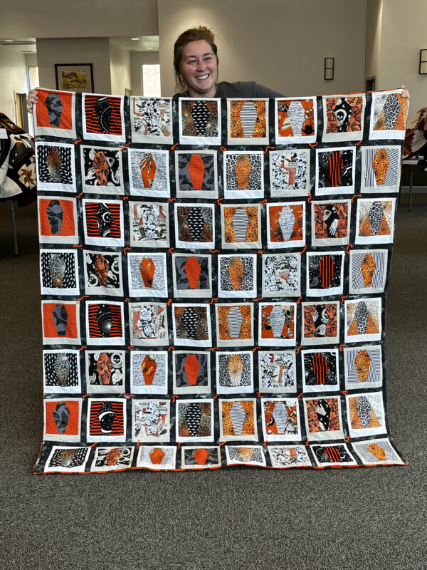 A woman holds up an intricate white, orange and black patterned quilt.
