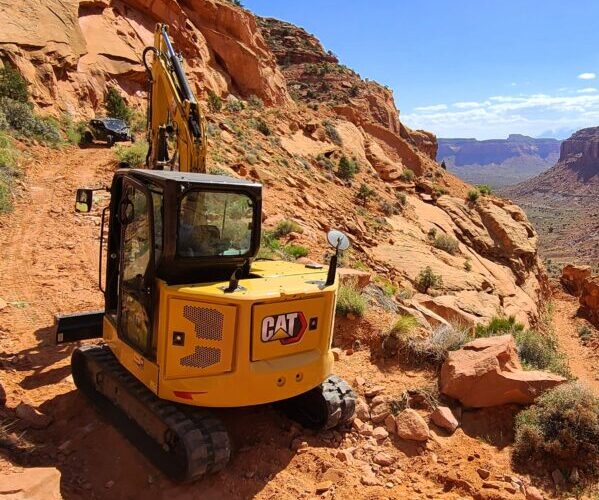 A backhoe scales steep red sand dirt road in the middle of the day.