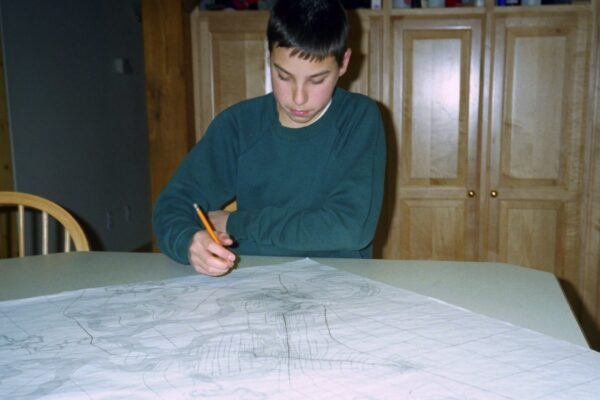 A teenage boy draws on a large map at the kitchen table.