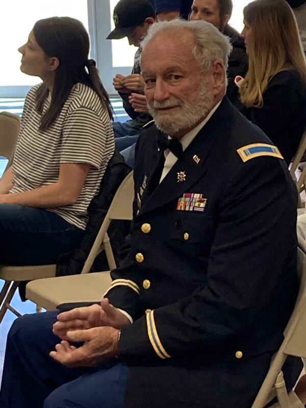 An elderly man sitting in a gym audience in a navy uniform.