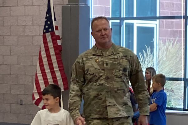 A man wearing an army uniform holds a boy's hand as they walk past rows of elementary school kids.