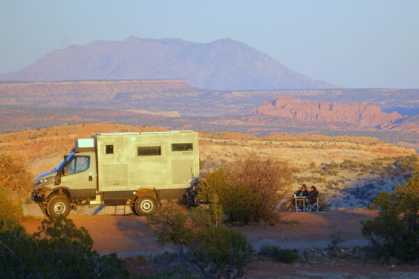 A van and dry campers on desert land in Escalante. You can see red and white mountains and a foggy blue sky in the background.