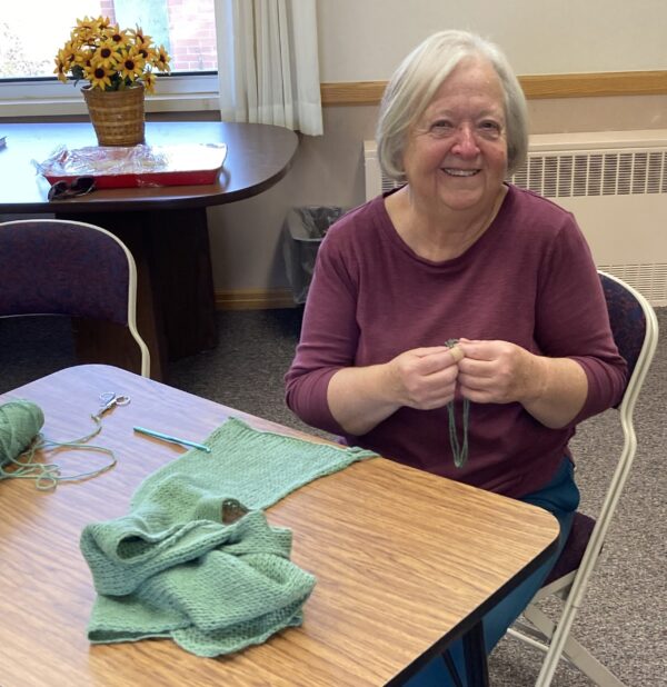 A woman sits at a table, preparing to put fringes on a green crocheted scarf.