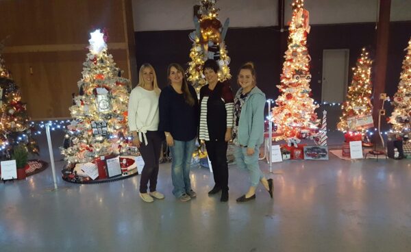 Four women stand in front of a line of lit trees at the Garfield County fair building for the Panguitch festival of trees.
