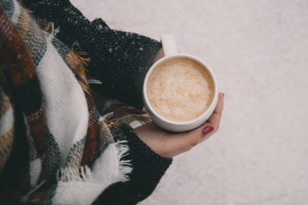 A mug of hot chocolate held by a person in a scarf. There is snow visible on the ground in the background.