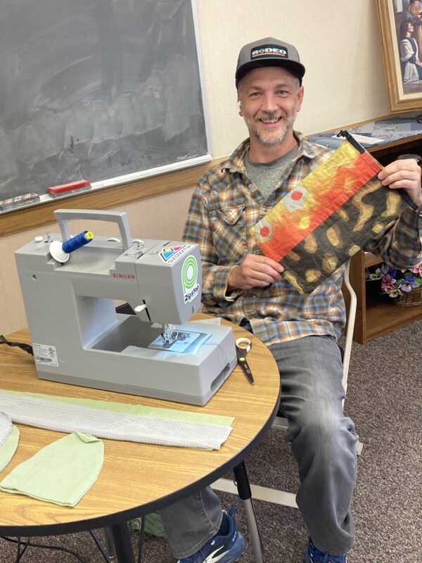 A man sits at a sewing machine, showing off his colorful work.
