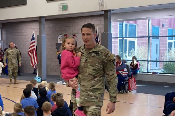A man in an army uniform visits an elementary school, carrying a small girl.