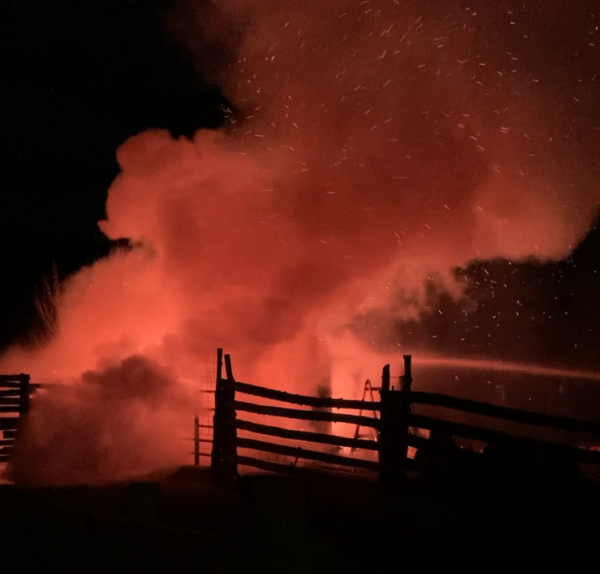 A billow of red smoke rises to the sky at night near a farmyard fence.