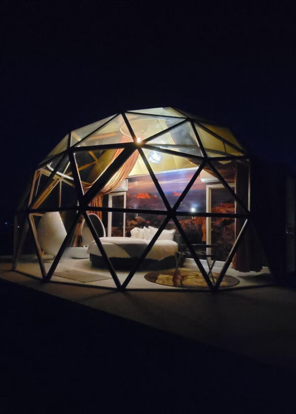A glass geodesic dome made up of triangular panels glows with light, surrounded by darkness at night. Curtains that slide all the way around are shown pulled to the side in the back.
