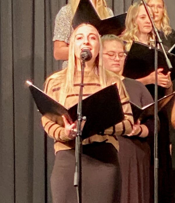 A high school girl holding a choir book sings into a microphone.