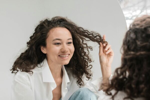 A young adult woman plays with her hair while looking in the mirror. She looks happy and content with what she sees.