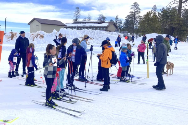 Children wearing thin cross-country skis line up on a graded snow path. It's a nice sunny day, but they're still dressed for the snow.
