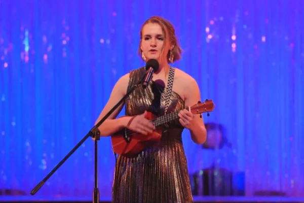 Clara, wearing dark makeup, dangly earrings, and a silvery-black dress with one shoulder, plays the ukulele and sings in a microphone onstage.