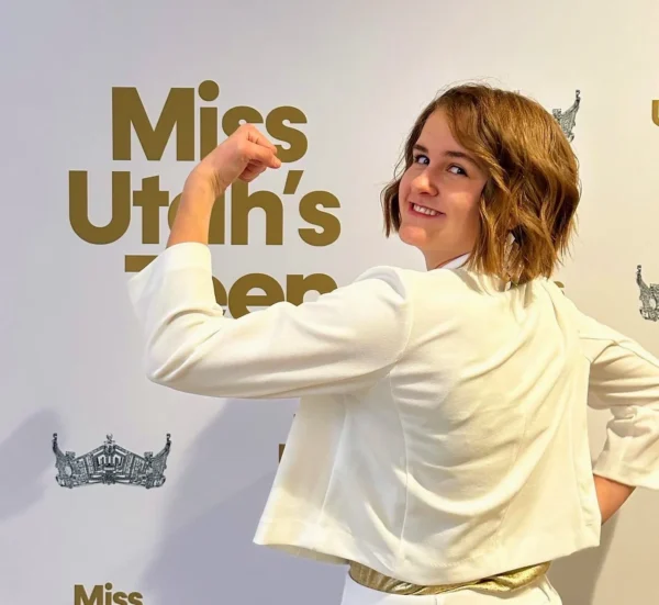 Clara does a showing-muscles pose, flexing her bicep. She stands backwards, facing the Miss Utah's Teen backdrop.
