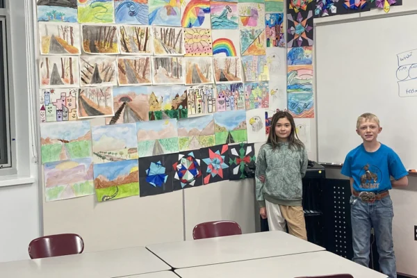 Two elementary school students stand by a wall of paper creations, rainbow drawings, and beginning one-point perspective artwork.