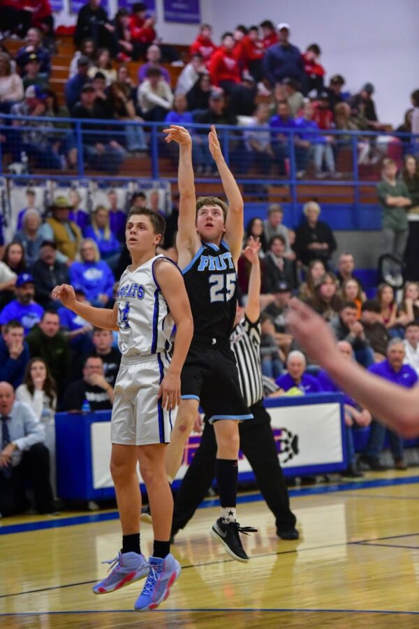 A Piute boys basketball player hangs in the air after taking a 3-point shot. A Panguitch player watches on anxiously.