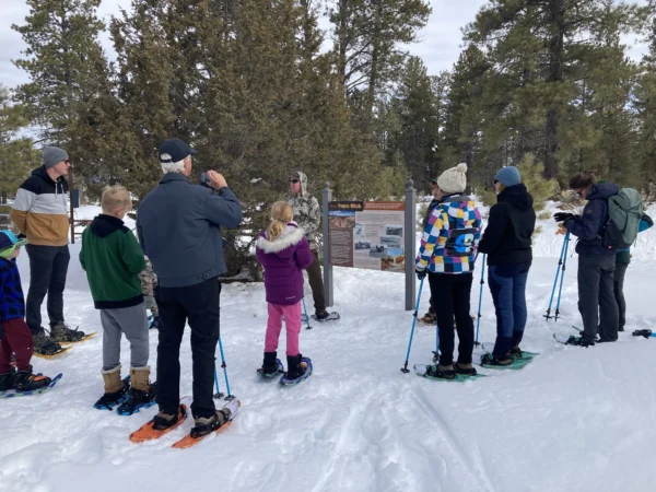 A group of people of all ages in snow shoes gathers around a park sign and listens to a guide.