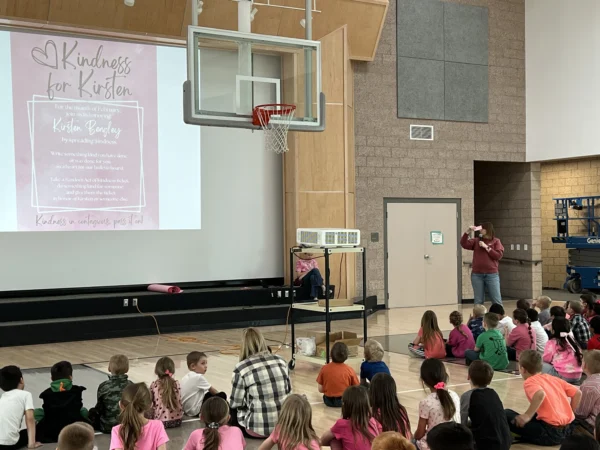 Kids sit in Bryce Valley Elementary School's gym, listening to Trista, who stands in the front talking near a projector. The projector shows a "kindness for Kirsten" flyer.