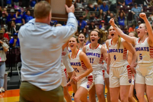 A bunch of girls basketball players cheer excitedly as they approach a man holding a trophy.