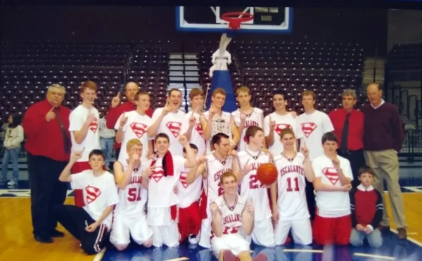 Group picture of 2008 Escalante boys basketball team in red and white uniforms and superman shirts. Coaches stand on either side.