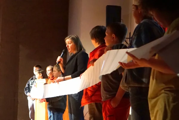 Janet Sumner Johnson speaking in a microphone on a stage. Elementary school kids stand their with her, holding a long string of white papers taped together.