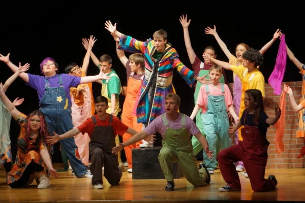 The cast of Joseph and the Amazing Technicolor Dreamcoat at Bryce Valley strikes a big ending pose. They wear colorful overalls and Joseph, in the middle, is wearing his colorful coat.