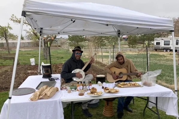 Two men play a guitar and a banjo while they sit at their booth at the farmer's market.