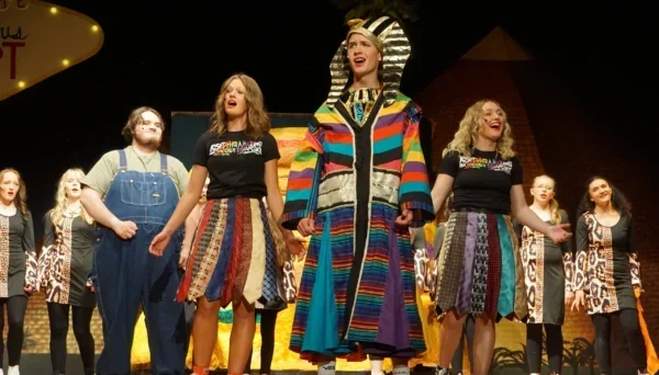 Joseph (Eli Beesley) in an Egyptian hat and necklace. He also wears his brightly colored Dreamcoat. The Narrators (Bradi Gates and Taylor LeFevre) and Jacob sing behind him.