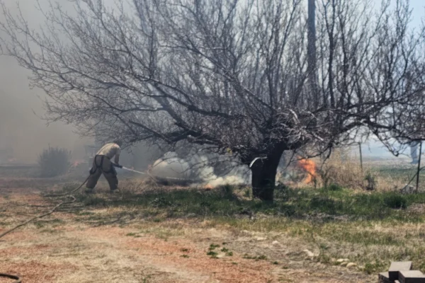 A man in a hazy cloud of smoke fights a fire away from a propane tank with a garden hose. He is not dressed like a firefighter.