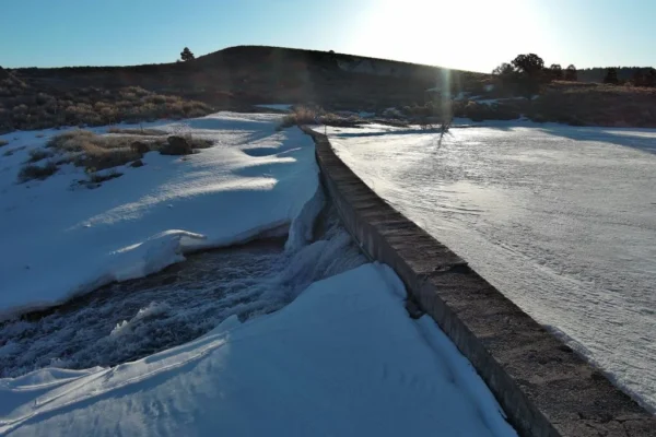 The concrete wall atop Panguitch Lake Dam bends as water runs out of a crack in the dam. The lake is iced over and there is snow everywhere.