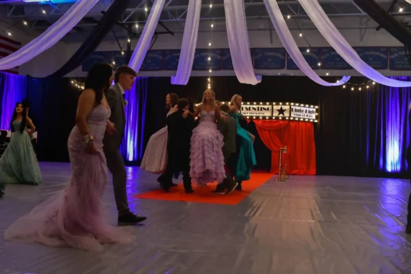 A circle of alternating boys and girls in tuxedos and formal dresses. The boys face inside the circle, lifting the girls, who sit facing outside.