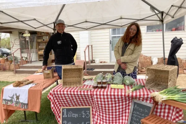 Bob sells bagels on an orange table cloth under the same tent canopy as Halfacre's Farm selling fresh vegetables on a gingham table cloth at the Escalante Farmer's Market.