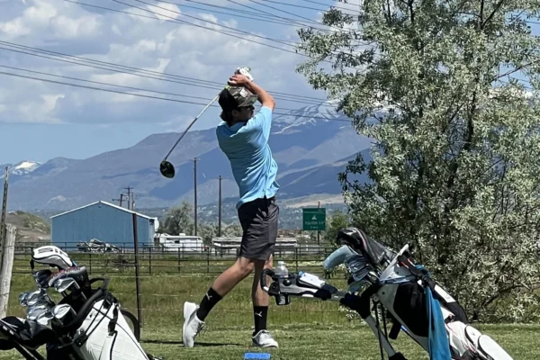 Swade Olsen swings his club on a golf course. Mountains are shown in the background of the course. In the foreground are a couple of golf bags.