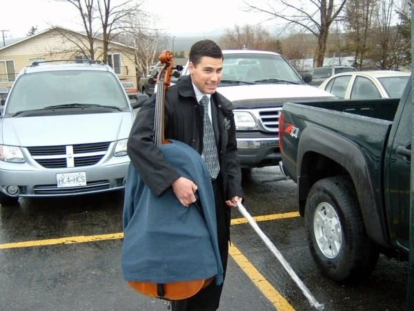 Elder Martel carries a cello inside from a rainy parking lot. The cello has a shirt over it.