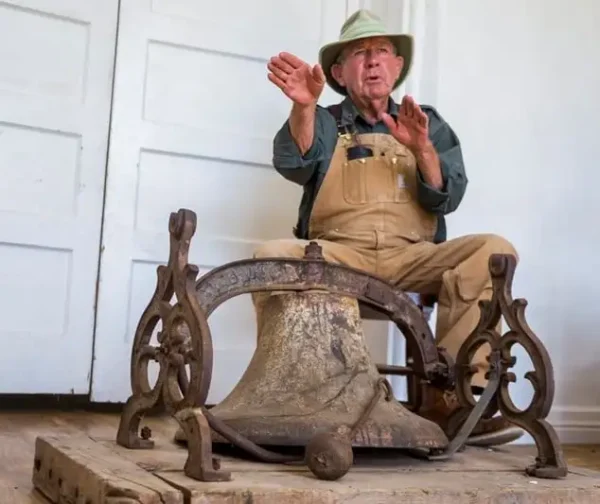 A man gestures to an old, rusty bell as he speaks.