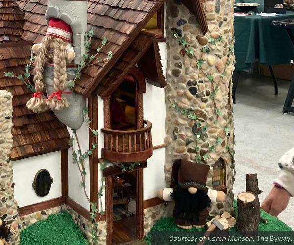 Two gnomes at their home. One looks out a tower window; the other stands outside by an ax, some logs and a chopping block. Some light can be seen through another window.