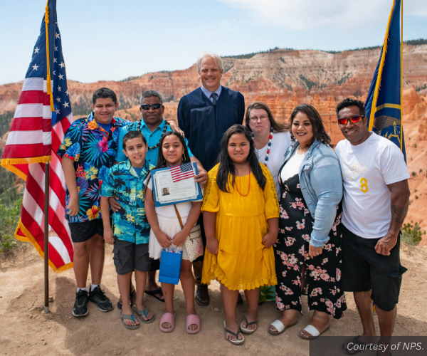 A large family stands with U.S. Magistrate Judge Dustin B. Pead between an American and a Utah flag. Behind them are the red hoodoos of the Bryce Canyon amphitheater. Courtesy of NPS.