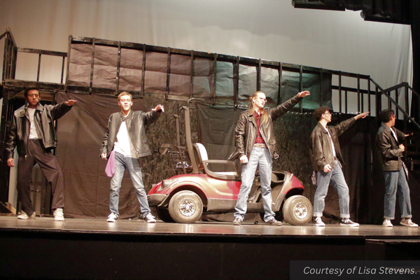 Five boys in leather jackets dance on stage with a car on set behind them. Courtesy of Lisa Stevens.