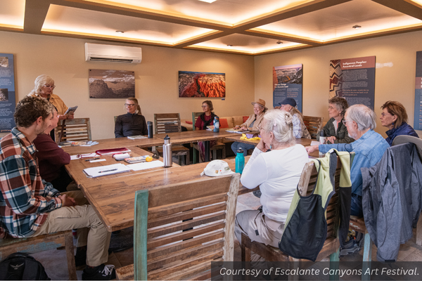 A number of participants sit at a square table listening to a presenter at an Escalante Art Festival workshop.