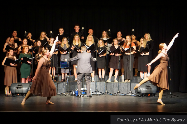 Tyrel Ivie leading the Piute choir in singing as two girls from Piute's dance company flit across the stage in front of them. Courtesy of AJ Martel, The Byway.