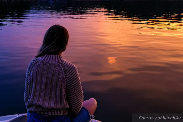 A girl sitting on a boat looks out over the water in a lake as the sun sets. Courtesy of Hitchhike.