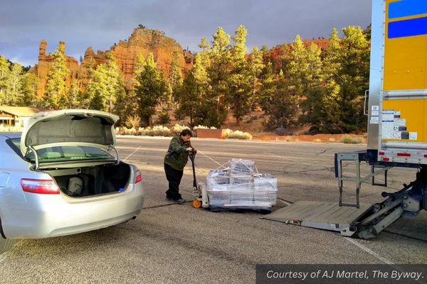 A man unloads stacks of newspapers off a semitruck and into the trunk of a silver sedan. Courtesy of AJ Martel, The Byway.